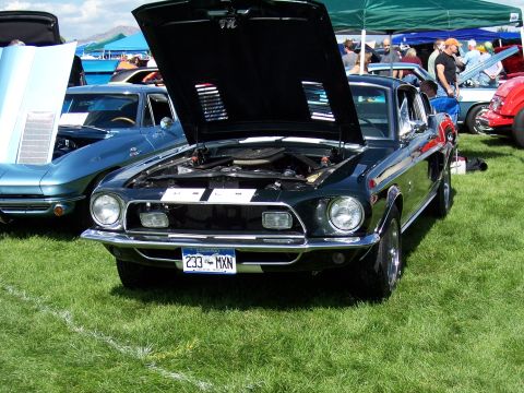 1972 Ford Ranchero - A dark green 1968 Ford Mustang is displayed with its hood open at an outdoor car show, parked next to a blue Chevrolet Corvette.