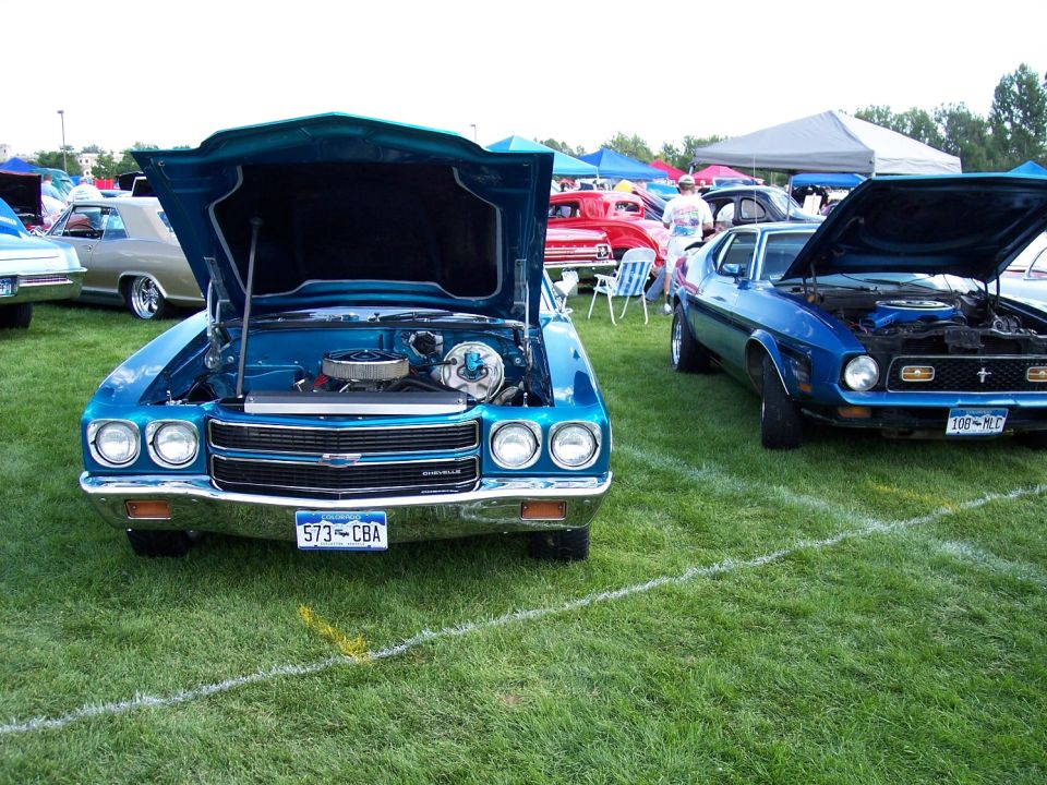 Two classic cars, a teal Chevelle and a blue Ford Mustang, displayed with hoods open at a car show