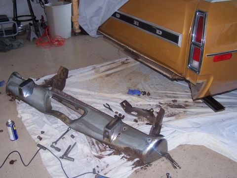 1973 Ford Ranchero - A detached chrome bumper lies on a white tarp next to the rear of a gold 1972 Ford Ranchero.