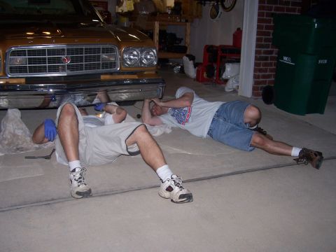 1973 Ford Ranchero - Two men are lying on the ground, working on the front end of a car in a garage.
