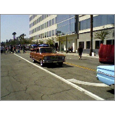 2007 Ford Ranchero - An orange pickup truck drives down a street with a crowd of people and buildings in the background.
