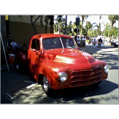 2007 Ford Ranchero - A bright red pickup truck is parked on a street, with people and trees in the background.