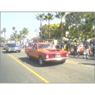 2007 Ford Ranchero - A red coupe utility vehicle with a hood scoop drives down a street lined with palm trees and spectators.