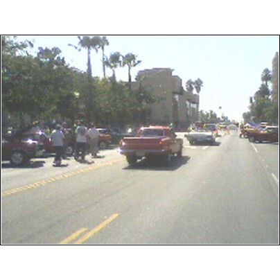 2007 Ford Ranchero - A red pickup truck drives down a street lined with trees and buildings, with several other vehicles present.