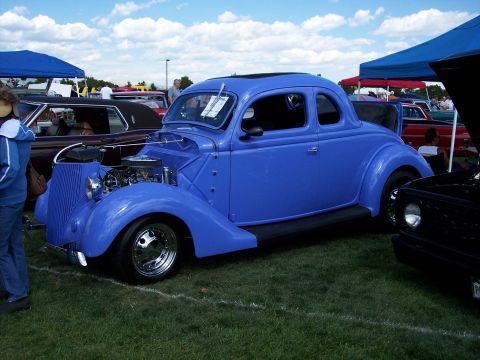1972 Ford Ranchero - A blue hot rod coupe is displayed at a car show, with other vehicles and tents visible in the background.