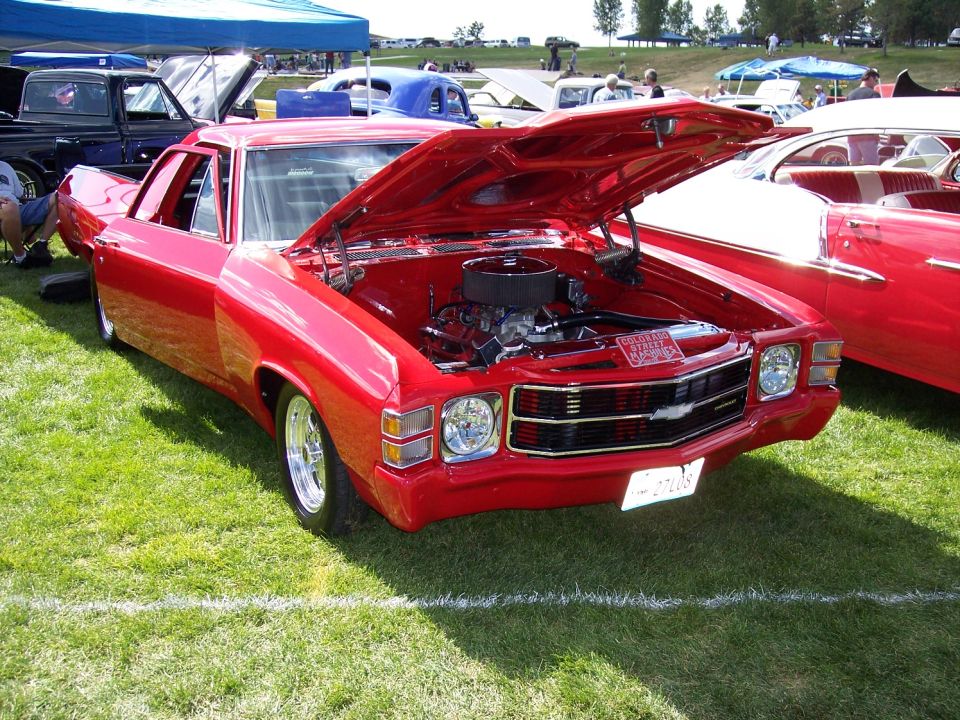 Red 1970s Chevrolet El Camino parked on grass, hood open showing engine. Another red car is parked to the right
