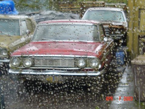 1964 Ford Fairlane - A red 1964 Ford Fairlane sedan is parked next to other vehicles, viewed through a window covered in raindrops.