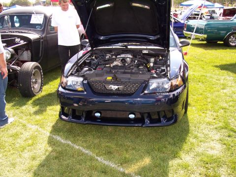 1972 Ford Ranchero - A dark blue Ford Mustang is displayed with its hood open, revealing the engine. The vehicle is parked on grass at an outdoor event.