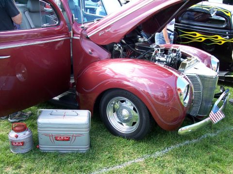 1972 Ford Ranchero - A custom red hot rod with flame pinstriping and a chrome grille is displayed with its hood open, next to another vehicle with flame graphic…