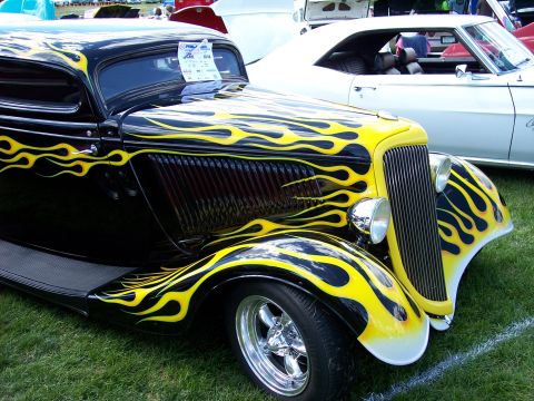 1972 Ford Ranchero - A black hot rod with vibrant yellow flame graphics is displayed on a grassy field, with another vehicle visible in the background.