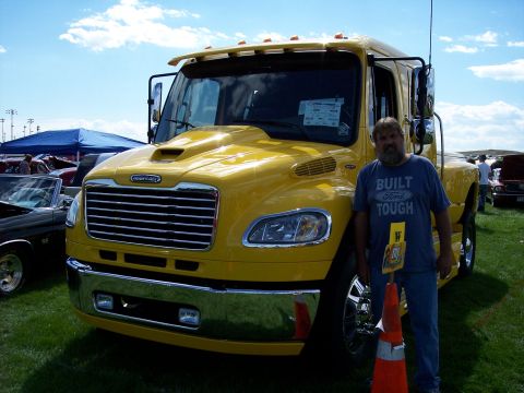 1972 Ford Ranchero - A man poses with a bright yellow Freightliner semi-truck at a car show, with other vehicles and tents in the background.