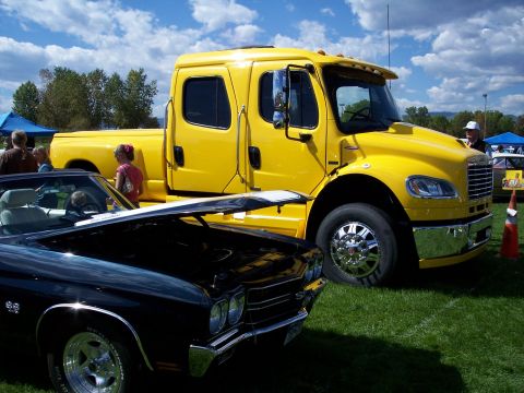 1972 Ford Ranchero - Two vehicles, a yellow Freightliner truck and a black Chevrolet Chevelle, are displayed on a grassy field under a cloudy sky.