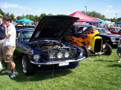 1972 Ford Ranchero - A blue 1969 muscle car is displayed with its hood open at an outdoor car show, with a hot rod flame graphics behind it.