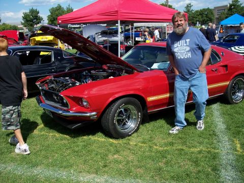 1972 Ford Ranchero - A red 1969 Ford Mustang Mach 1 is displayed with its hood open at an outdoor car show, alongside a man wearing a Ford-themed t-shirt.