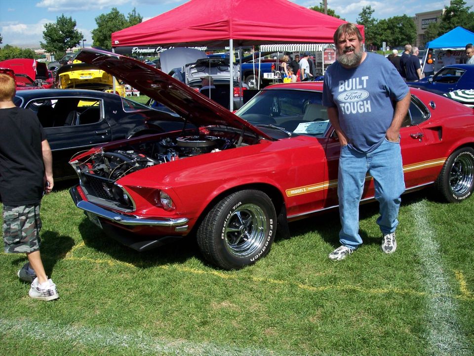 Red 1969 Ford Mustang Mach 1 with hood open, showing engine. Man in 'Built Ford Tough' shirt stands beside it