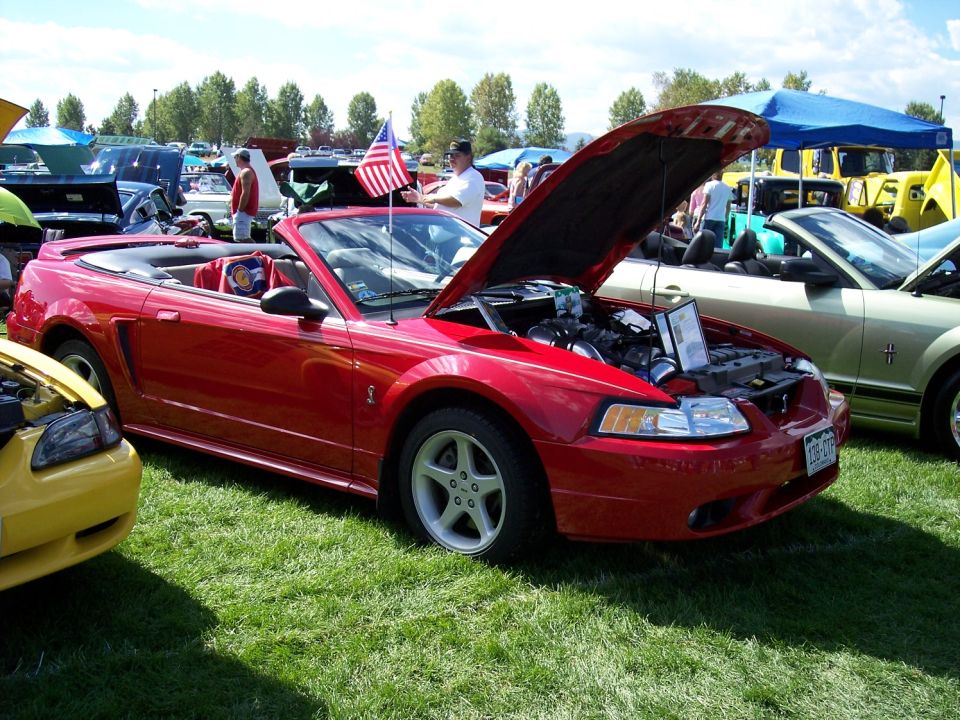 Red convertible sports car with hood open, American flag on antenna, parked on grass at car show