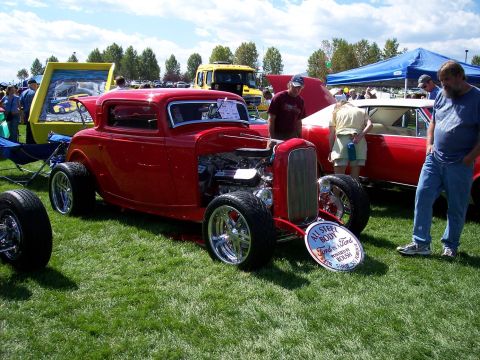 1972 Ford Ranchero - A red hot rod coupe is displayed on grass with other vehicles and people around.