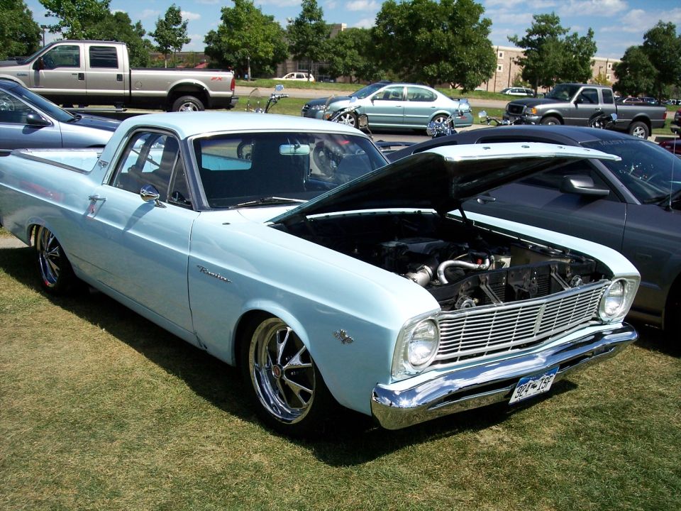 Light blue Ford Ranchero with hood open, showing engine. Chrome wheels. Other vehicles visible in background