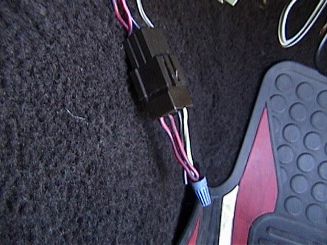 Black electrical connector with pink, white, and blue wires attached to a blue wire nut, on dark carpet next to a red and black floor mat