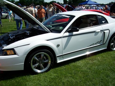 1972 Ford Ranchero - White Ford Mustang with black racing stripes and "40th Anniversary" decal is displayed with its hood open at an outdoor car show.