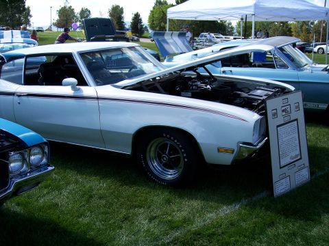 1972 Ford Ranchero - White coupe on display at an outdoor car show, hood open, with a descriptive board nearby.