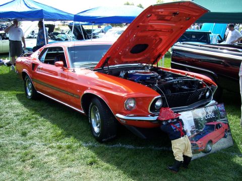 1972 Ford Ranchero - A child inspects the engine of a bright orange 1969 Ford Mustang Mach 1 at a car show.