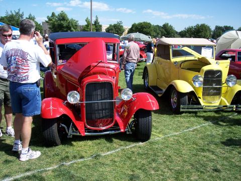 1972 Ford Ranchero - A bright red hot rod with its hood raised is displayed at an outdoor car show, with a yellow vintage car visible to its right.