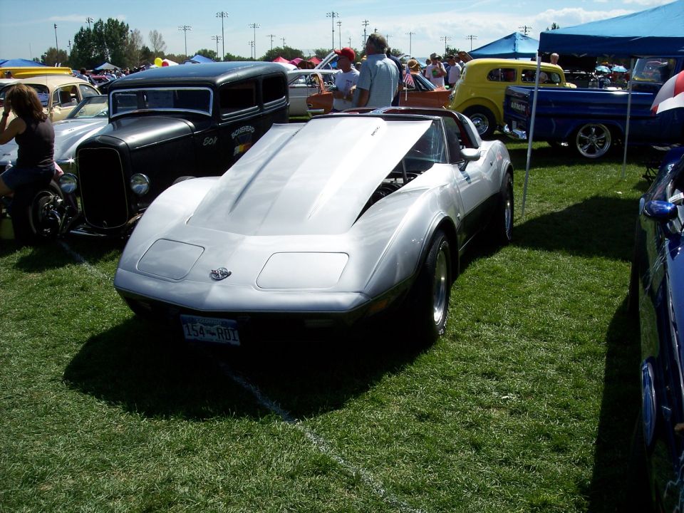 Silver 1975 Chevrolet Corvette C3 with hood open, parked on grass at car show. License plate reads 154-RDI
