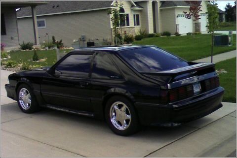 1989 Ford Mustang - A black 1989 Ford Mustang coupe is parked on a concrete driveway in front of a suburban house.