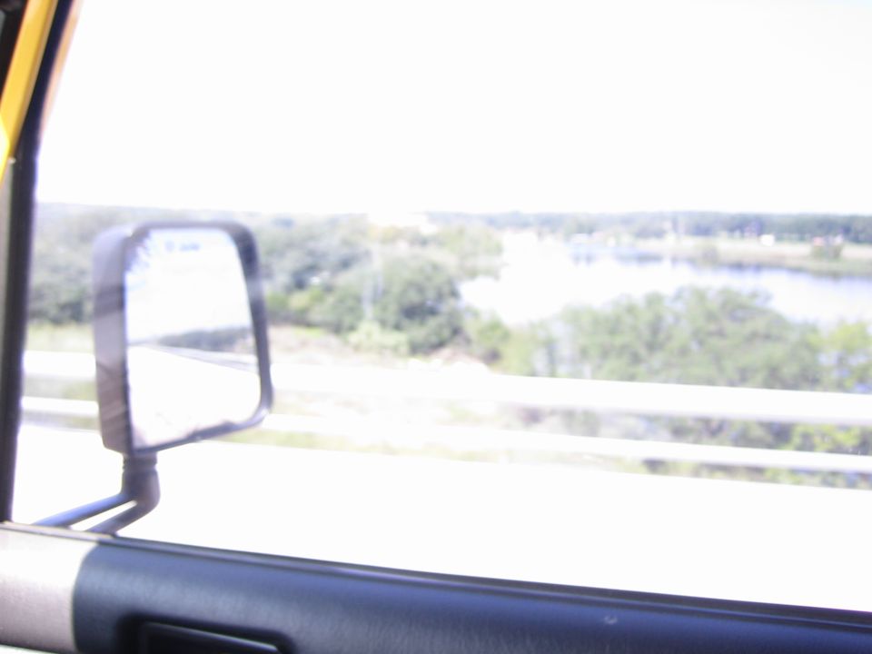 Yellow vehicle interior with side mirror reflecting a road and a distant body of water with trees