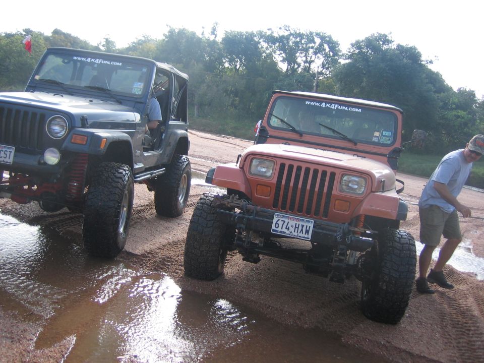 Two modified Jeep Wranglers, one dark blue and one orange, on a muddy dirt road. The orange Jeep has license plate 674-HXW