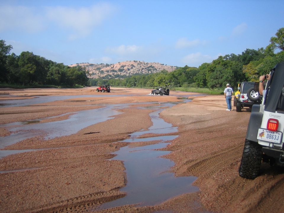 Several Jeeps driving on a sandy, wet riverbed with trees and hills in the background