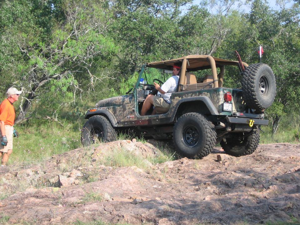 Man driving a camouflage Jeep Wrangler on a rocky trail. Another man stands nearby
