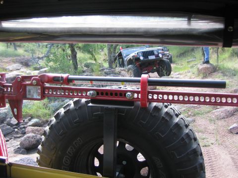 2001 Jeep Wrangler - A red hi-lift jack is mounted to the spare tire of a 2001 Jeep Wrangler, with another Jeep visible on a rocky trail.