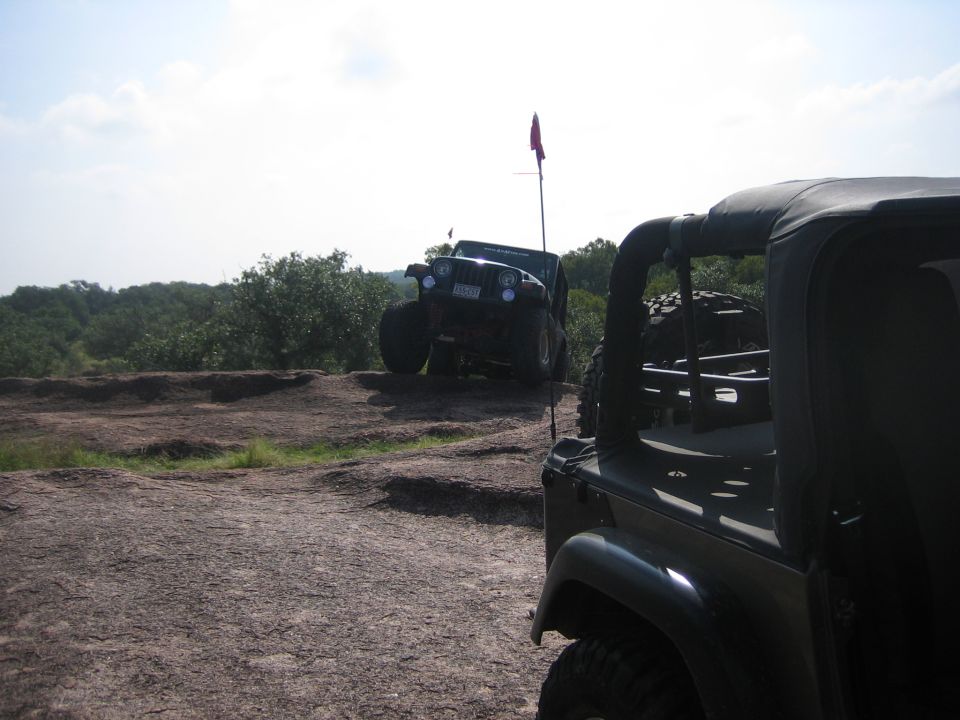 Dark green Jeep Wrangler in foreground, another Jeep Wrangler on rocky hill with red flag