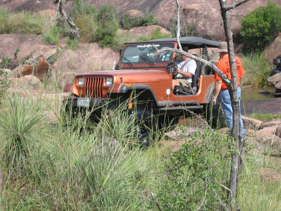 Orange Jeep Wrangler with soft top on rocky, grassy trail. Driver visible