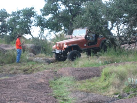 2001 Jeep Wrangler - A man stands near an orange Jeep Wrangler parked on a rocky, grassy landscape with trees in the background.