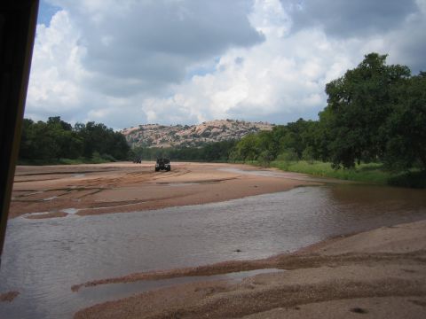 2001 Jeep Wrangler - Off-road vehicles traverse a sandy riverbed under a cloudy sky, with trees and hills ahead.