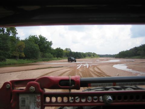 2001 Jeep Wrangler - View from inside a vehicle shows a black Jeep Wrangler driving on a sandy riverbed with trees lining the banks.