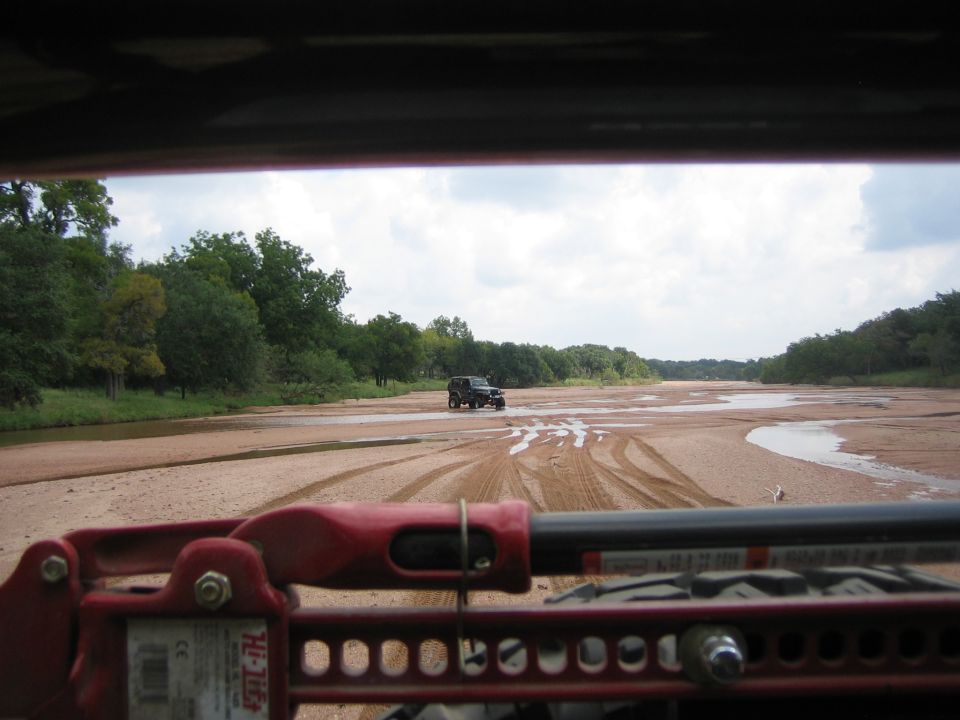 Red Hi-Lift jack and vehicle interior in foreground, black Jeep Wrangler driving on sandy riverbed