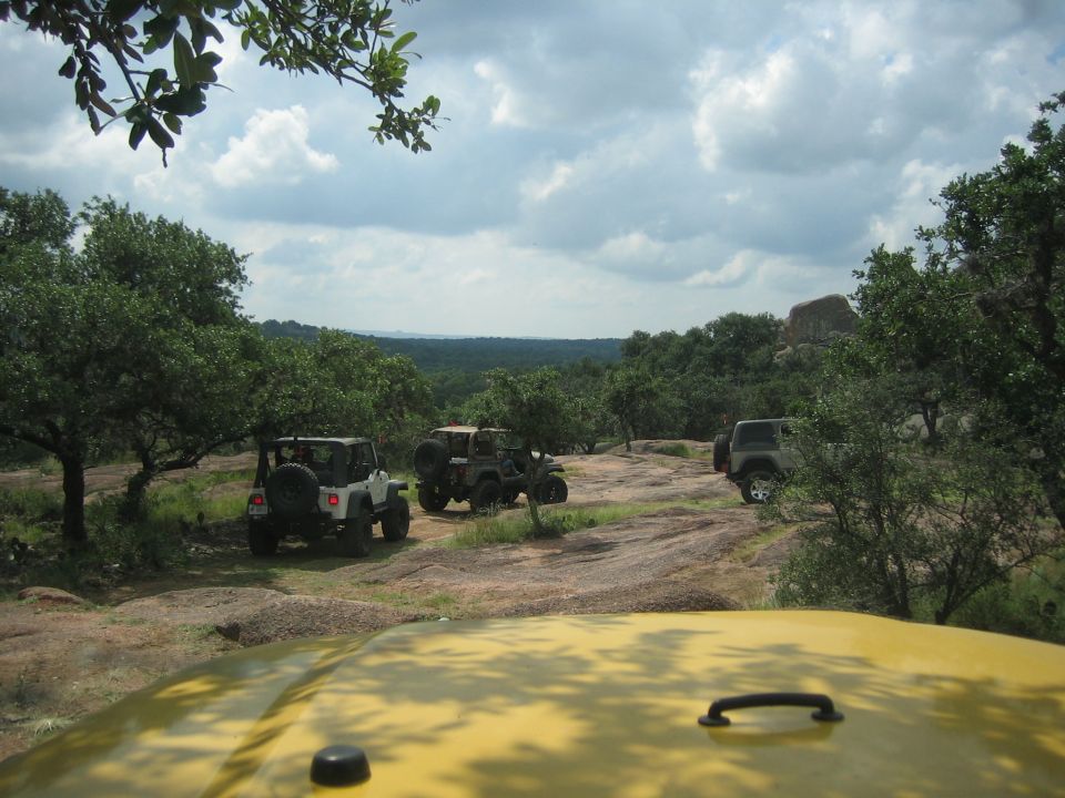 Yellow Jeep hood in foreground, three other Jeep Wranglers on a rocky, tree-lined landscape under a cloudy sky