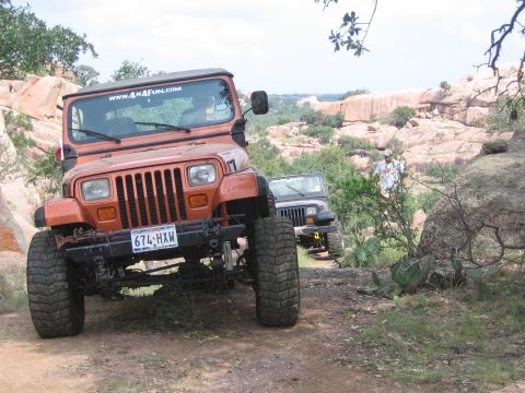 2001 Jeep Wrangler - An orange Jeep Wrangler is parked on a dirt path with another Jeep in the background, set against a rocky, natural landscape.