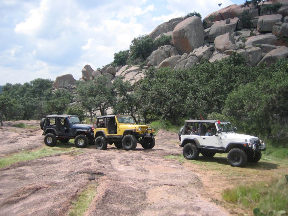 Three Jeep Wranglers parked on a rocky, wooded hillside. The yellow and blue Wranglers are side-by-side, with a white one parked slightly ahead and to the right