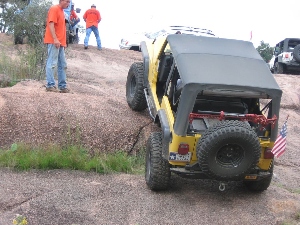 Yellow Jeep Wrangler climbing a rock face, spare tire on back, American flag attached