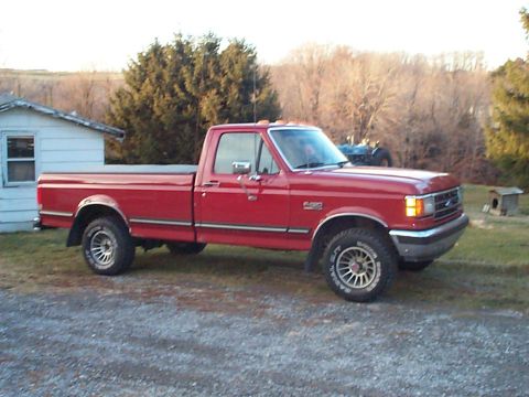 1989 Ford F-150 - A red Ford F-150 pickup truck is parked in a grassy area next to a small building.