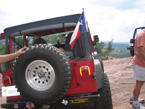 2001 Jeep Wrangler - Rear view of a red Jeep Wrangler flying a Texas flag, with a spare tire mounted. The vehicle appears to be a 2001 model.