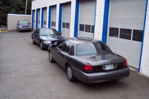 2001 Ford Crown Victoria - A dark grey 2001 Ford Crown Victoria is parked in front of a building with multiple garage doors, alongside a blue sedan and a dark SUV.