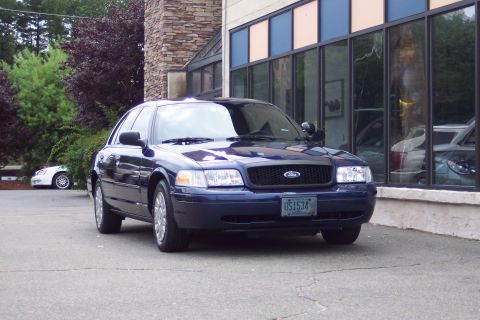 2001 Ford Crown Victoria - A dark blue 2001 Ford Crown Victoria police car is parked in front of a building with large windows. Another white vehicle is visible in th…