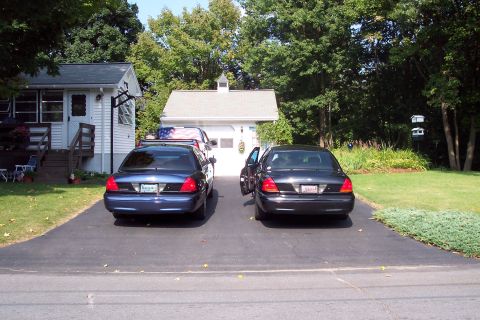 2001 Ford Crown Victoria - Two Ford Crown Victorias, a blue and a black model, are parked in a driveway in front of a garage.