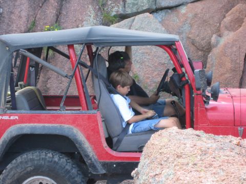 2001 Jeep Wrangler - Red Jeep Wrangler with soft top navigating rocky terrain, an adult driver and young child passenger visible inside the open cab.